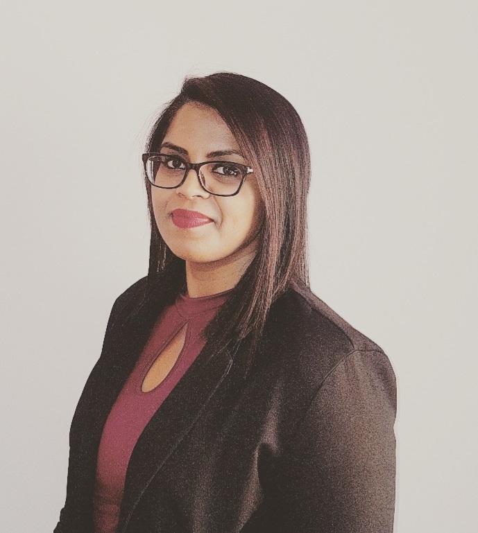 A person with glasses, wearing a blazer and maroon top, smiles slightly.