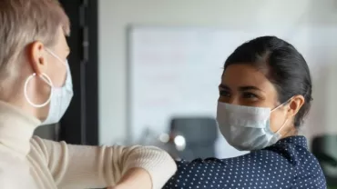 Two people in masks greet each other with elbow bump indoors.