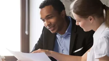 Two people reviewing a document at a desk with a laptop.
