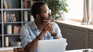 Person sitting at a desk, looking thoughtfully out a window, with a laptop and books in the background.
