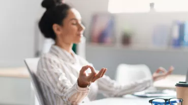 Person meditating at a desk with a laptop and glasses nearby.