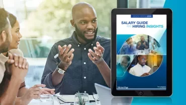 Person discussing with colleagues beside a tablet displaying a salary guide.