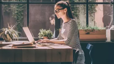 Person working on a laptop at a wooden table, plants in background.