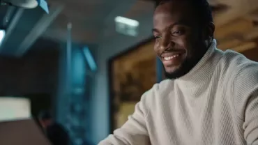 Person wearing a sweater, smiling while using a laptop in a modern office.