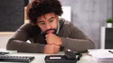 Person at a desk looking thoughtfully at a phone.