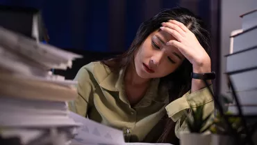 A stressed woman in a green shirt with a hand on her head in a dark office with piles of papers and books on either side of her