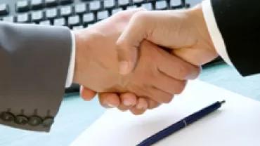 Two people shaking hands over a desk with a pen and keyboard visible.