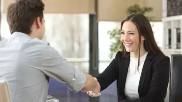Two people shaking hands at an office desk, smiling.