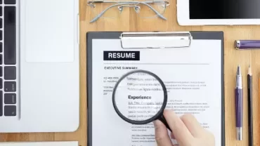 Hand holding magnifying glass over resume, next to laptop and office supplies.