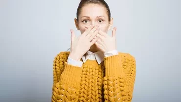 Person with hands covering mouth, wearing a mustard sweater.