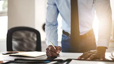 Person standing at a desk writing, sunlight behind.