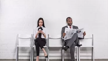 Two people sitting on chairs; one reads a newspaper, the other uses a phone.