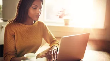 Person working on a laptop in a sunlit room with a mug in hand.