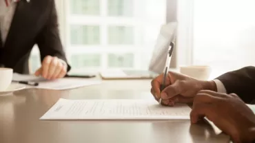 Person writing on a contract at a desk with a laptop and coffee cup.