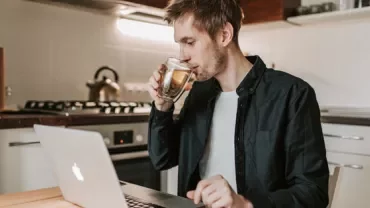Person drinking tea while using a laptop in a kitchen.