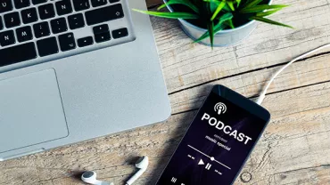 Smartphone on table playing podcast near laptop and earphones.