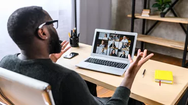 Person in a virtual meeting on a laptop, with shelves and books in the background.