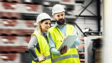 Two people in safety vests and helmets discuss plans on a tablet in a warehouse.