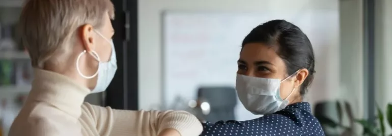 Two people in masks greet each other with elbow bump indoors.