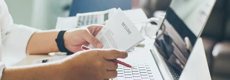 Person reviewing a resume at a desk with a laptop.