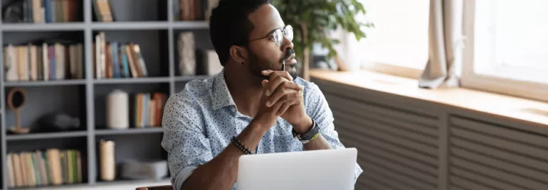 Person sitting at a desk, looking thoughtfully out a window, with a laptop and books in the background.