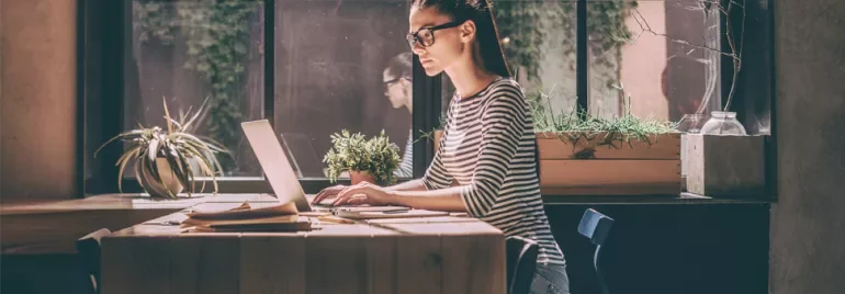Person working on a laptop at a wooden table, plants in background.