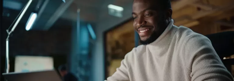 Person wearing a sweater, smiling while using a laptop in a modern office.