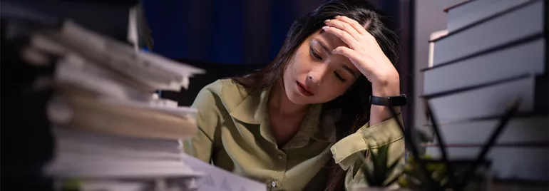 A stressed woman in a green shirt with a hand on her head in a dark office with piles of papers and books on either side of her