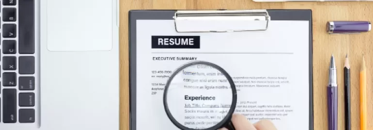 Hand holding magnifying glass over resume, next to laptop and office supplies.