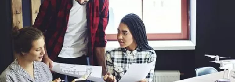Three people discussing and reviewing paperwork at a desk near a window.