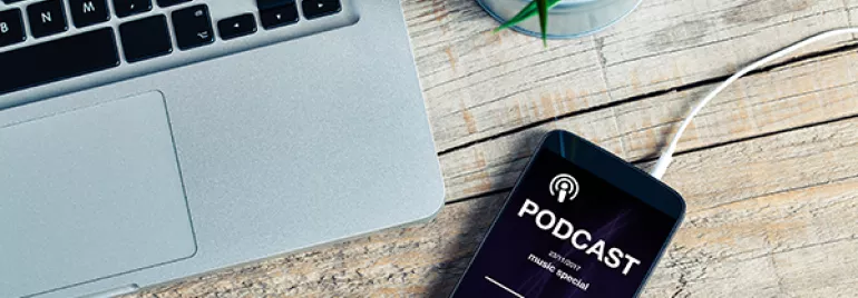Smartphone on table playing podcast near laptop and earphones.