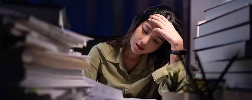 A stressed woman in a green shirt with a hand on her head in a dark office with piles of papers and books on either side of her