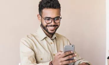 Person smiling at a smartphone with a laptop in the background.
