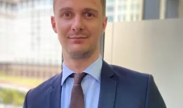 Person in a suit standing by a window with city buildings in the background.