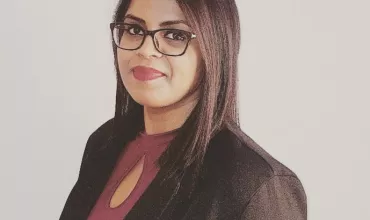 A person with glasses, wearing a blazer and maroon top, smiles slightly.