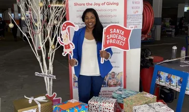 Person standing with Santa Shoebox signs near colorful gift boxes.