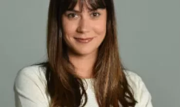 Personne souriante avec des cheveux longs et une chemise blanche sur fond gris.