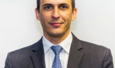 Person in a grey suit with a blue tie, smiling in front of a white background.