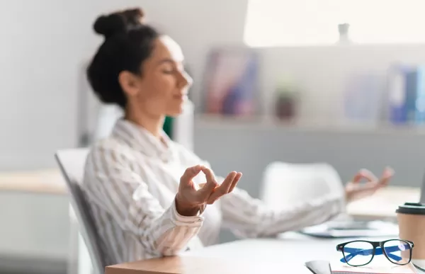 Person meditating at a desk with a laptop and glasses nearby.