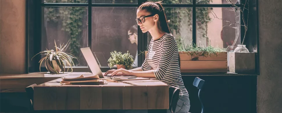 Person working on a laptop at a wooden table, plants in background.