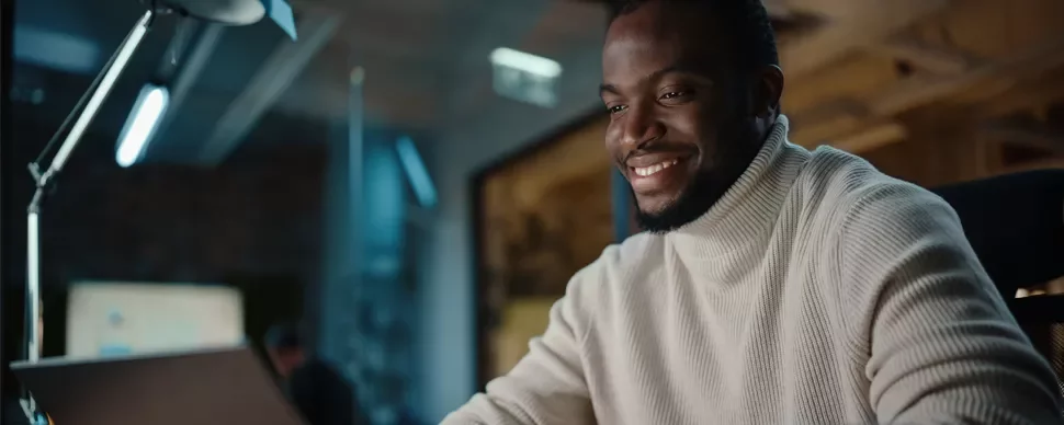 Person wearing a sweater, smiling while using a laptop in a modern office.