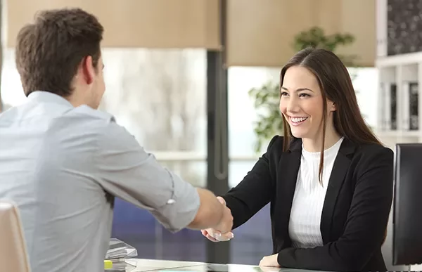 Two people shaking hands at an office desk, smiling.