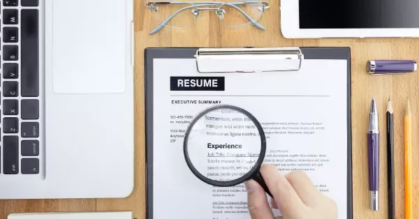 Hand holding magnifying glass over resume, next to laptop and office supplies.