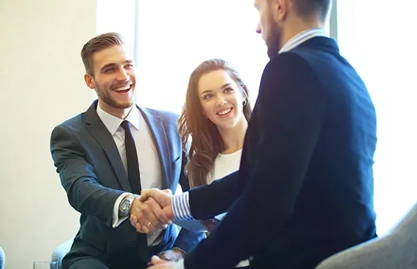 Three people in business attire, two shaking hands, smiling.
