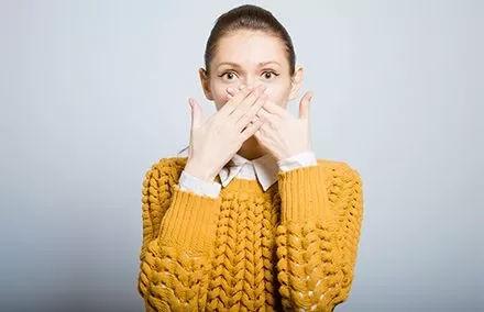 Person with hands covering mouth, wearing a mustard sweater.
