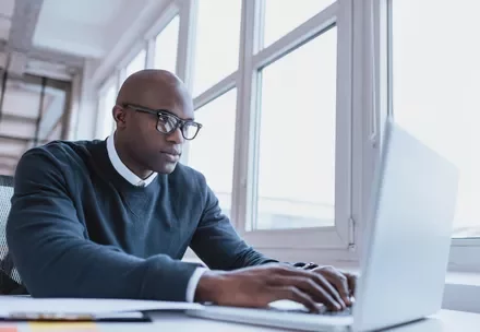 Person typing on a laptop at a desk near large windows.