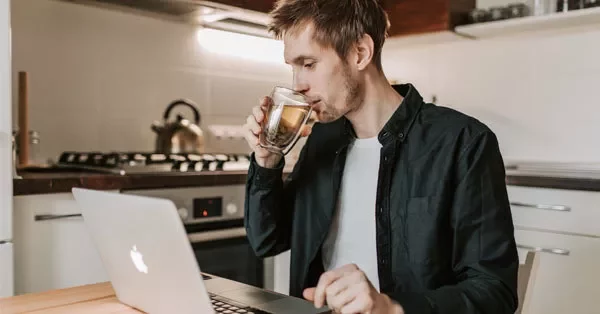 Person drinking tea while using a laptop in a kitchen.