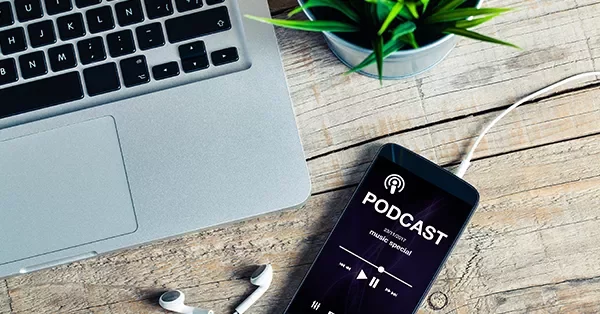 Smartphone on table playing podcast near laptop and earphones.