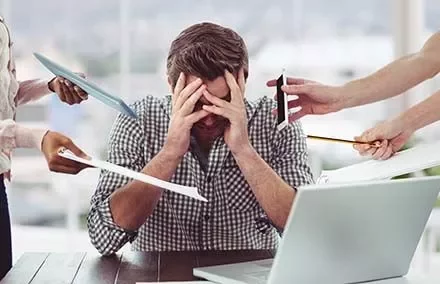 Person sitting at a desk overwhelmed with work, surrounded by papers and technology.